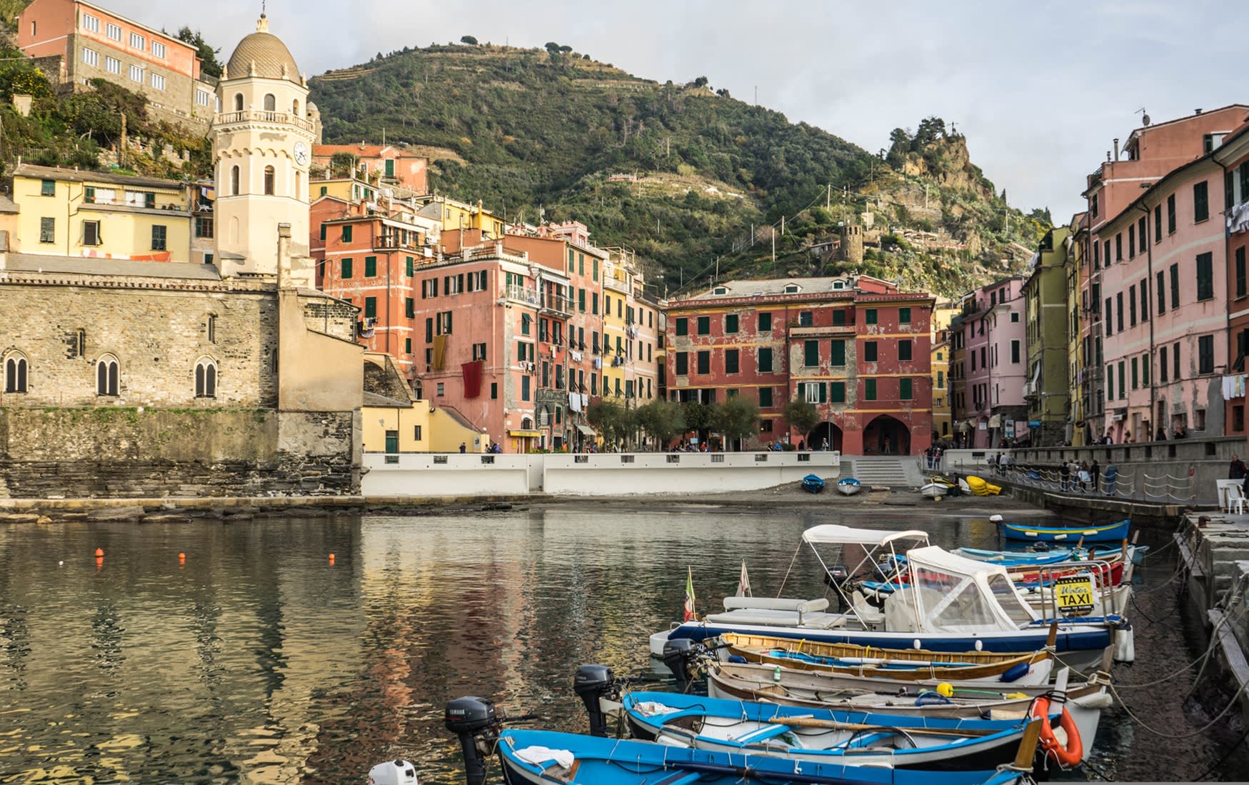Colorful buildings of Vernazza, Cinque Terre, Italy, with boats docked in the harbor and a green hillside behind.