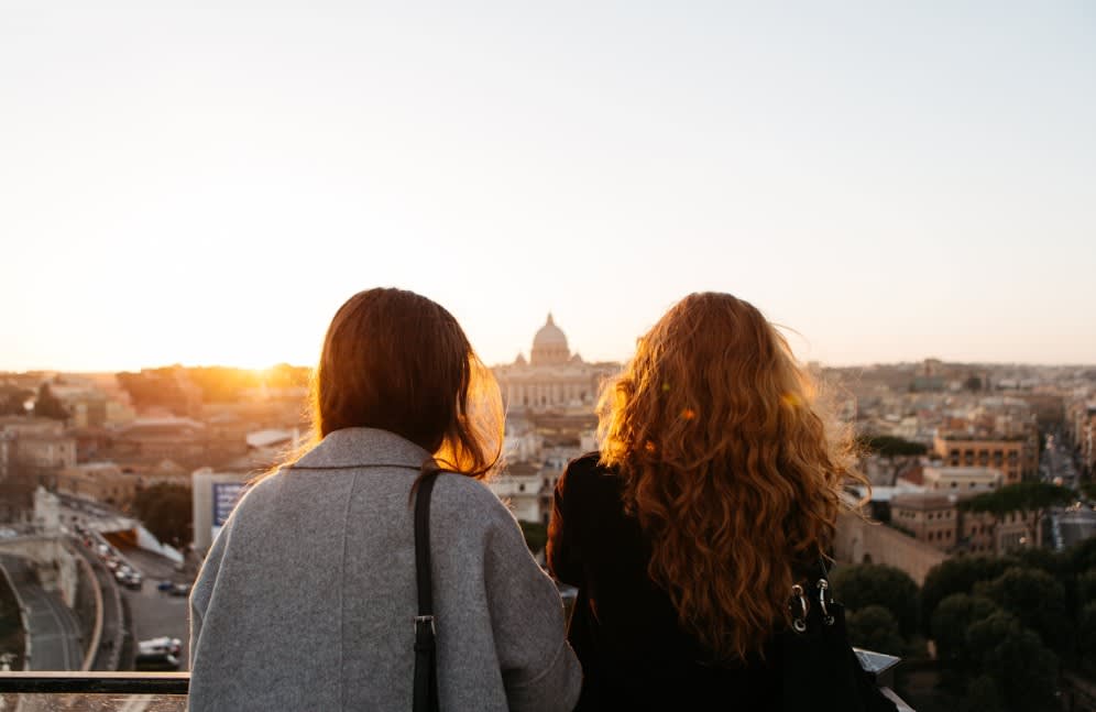 Two women overlooking Rome's skyline at sunset, with St. Peter's Basilica dome visible in the golden light.