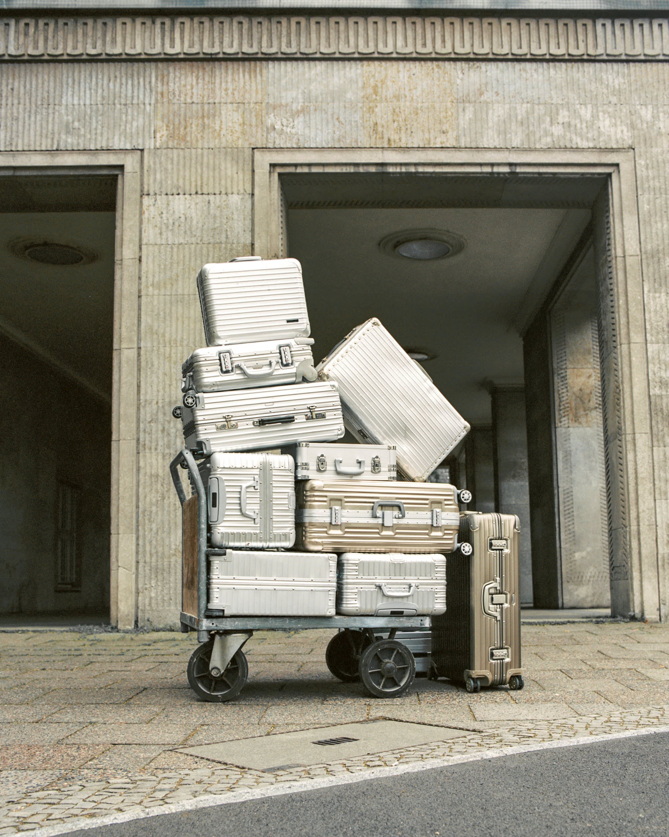 Stack of silver and gold aluminum hard-shell suitcases piled high on a luggage cart outside a stone building.