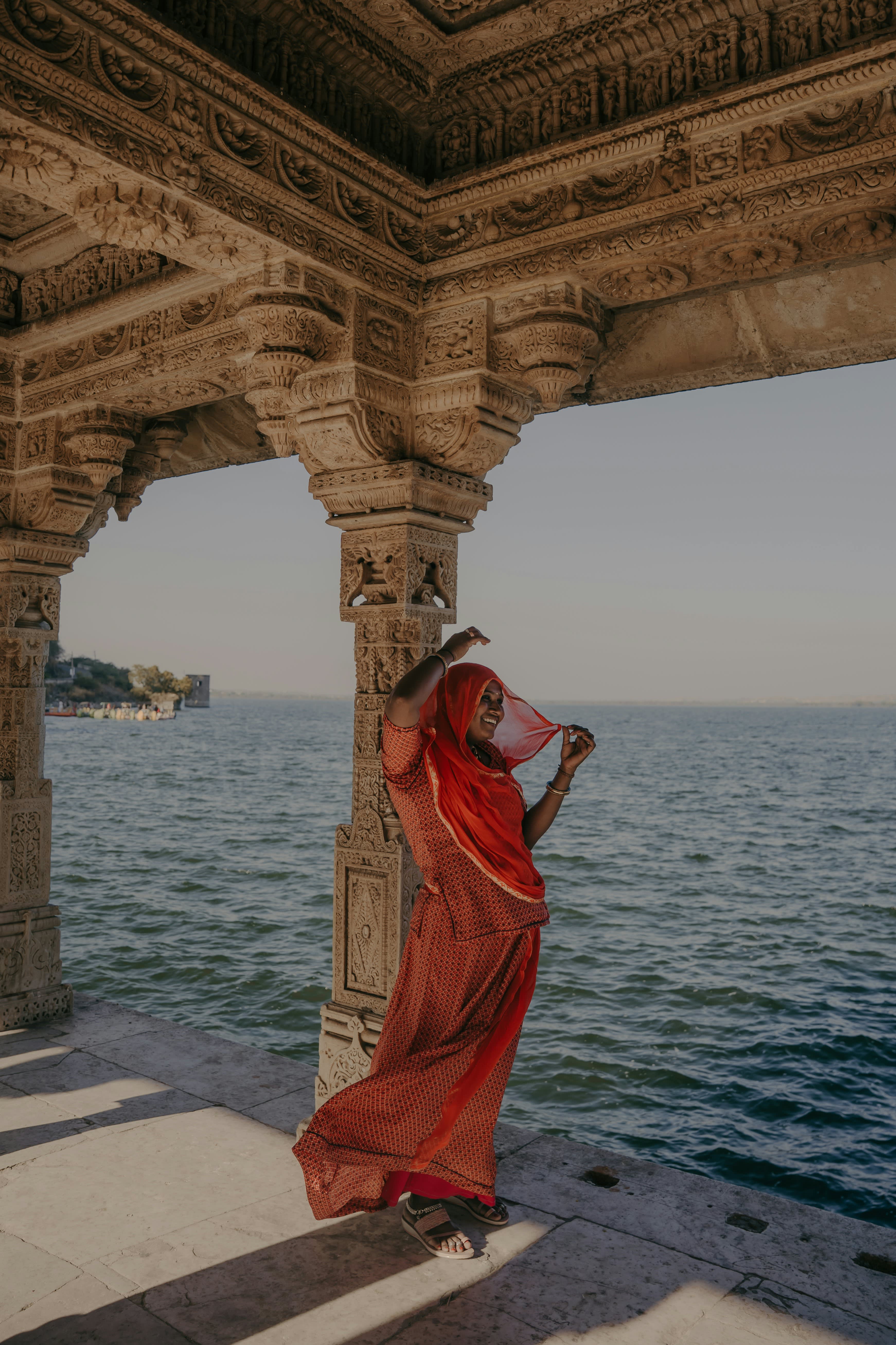 Woman in red saree posing beside ornate carved stone columns of an Indian temple overlooking a lake.