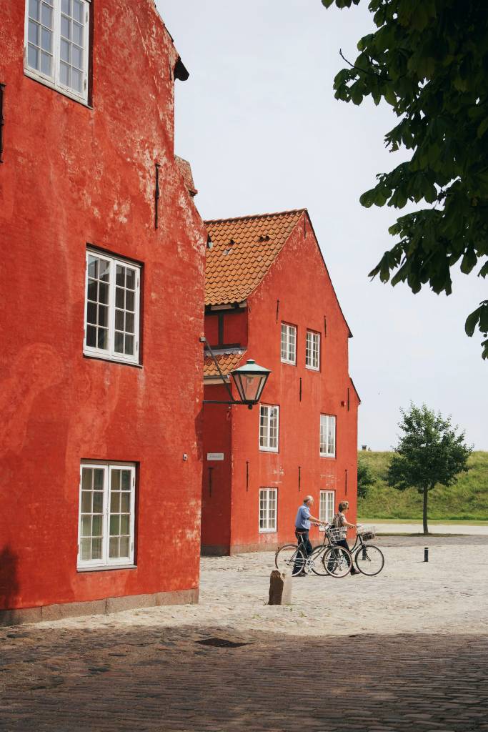 Two people with bicycles on a cobblestone square beside a large red historic building with white windows and a street lamp.