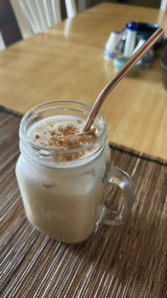 Creamy protein shake in a glass mason jar mug, topped with cinnamon and a rose gold metal straw, on a woven placemat.