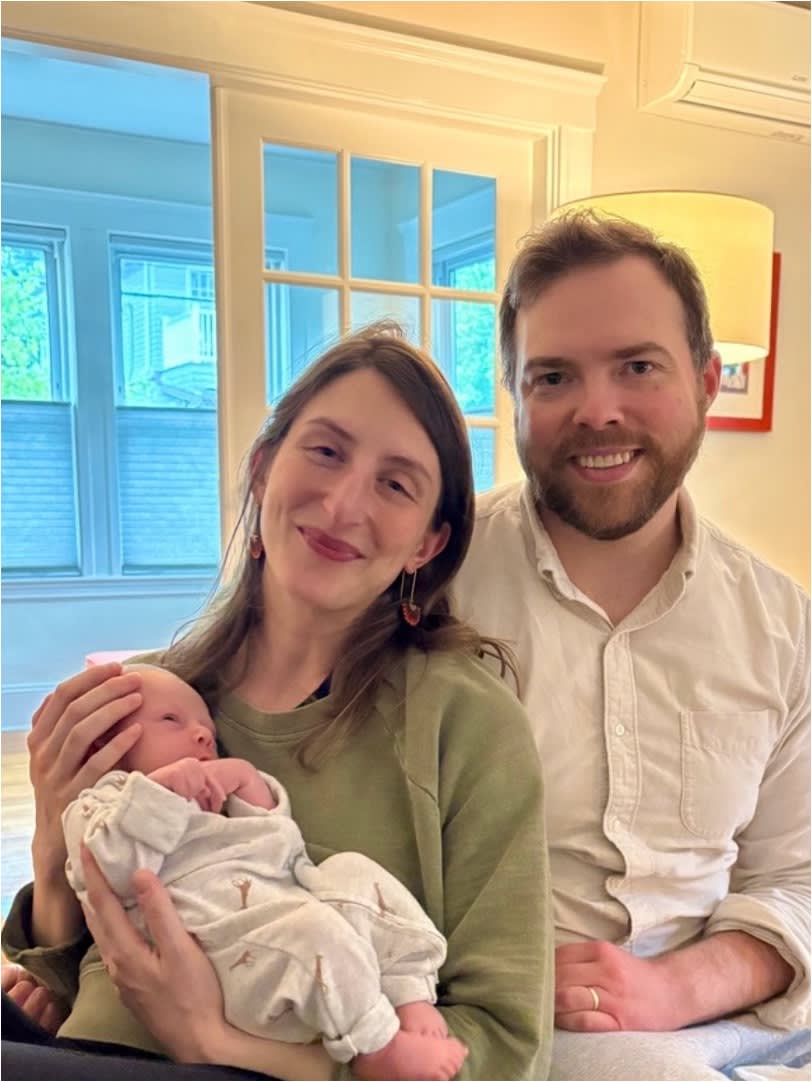 Smiling couple sitting together indoors, woman holding a newborn baby dressed in white.