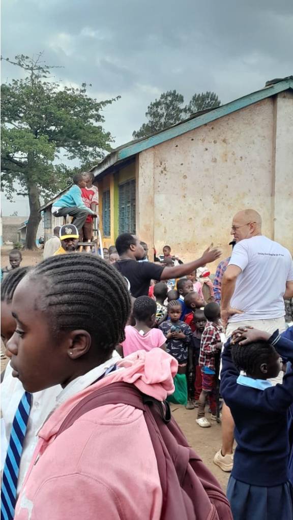 A large group of children and adults gathered outside a weathered building in Africa, with a bald man in a white shirt interacting with the crowd.