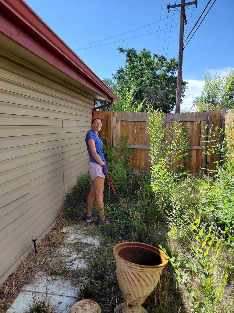 Smiling volunteer in blue shirt and cap holding a rake, clearing overgrown weeds beside a house on a sunny day.
