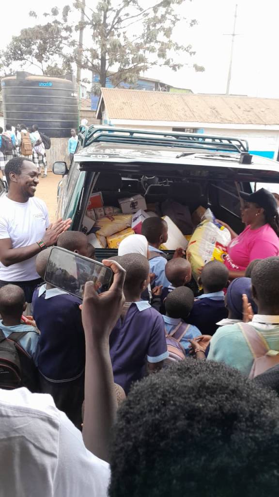 Volunteers unloading food donations from a vehicle, surrounded by excited schoolchildren in uniforms during an ETAfrica community outreach day.