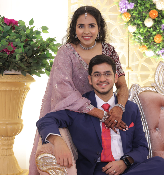 South Asian wedding couple; woman in pink dupatta with mehndi stands behind seated man in navy suit and red tie, surrounded by flowers.