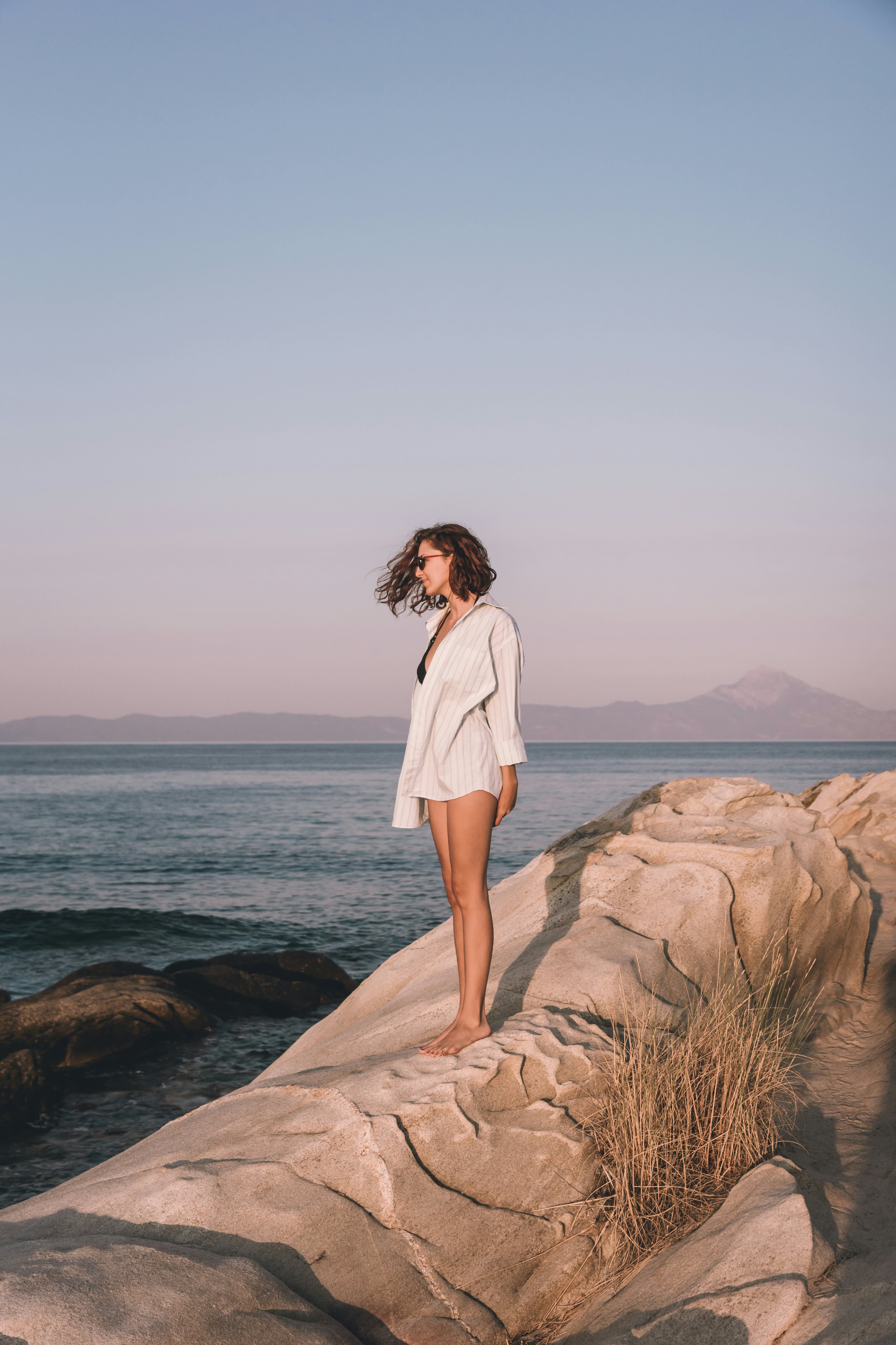 Woman in white shirt standing barefoot on coastal rocks, hair blowing in wind, calm sea and mountain silhouette at dusk behind her.
