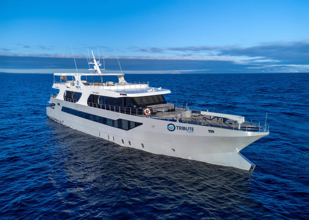 White luxury motor yacht "Tribute by Oniric" sailing on calm blue ocean under clear sky, multiple decks with open-air areas.