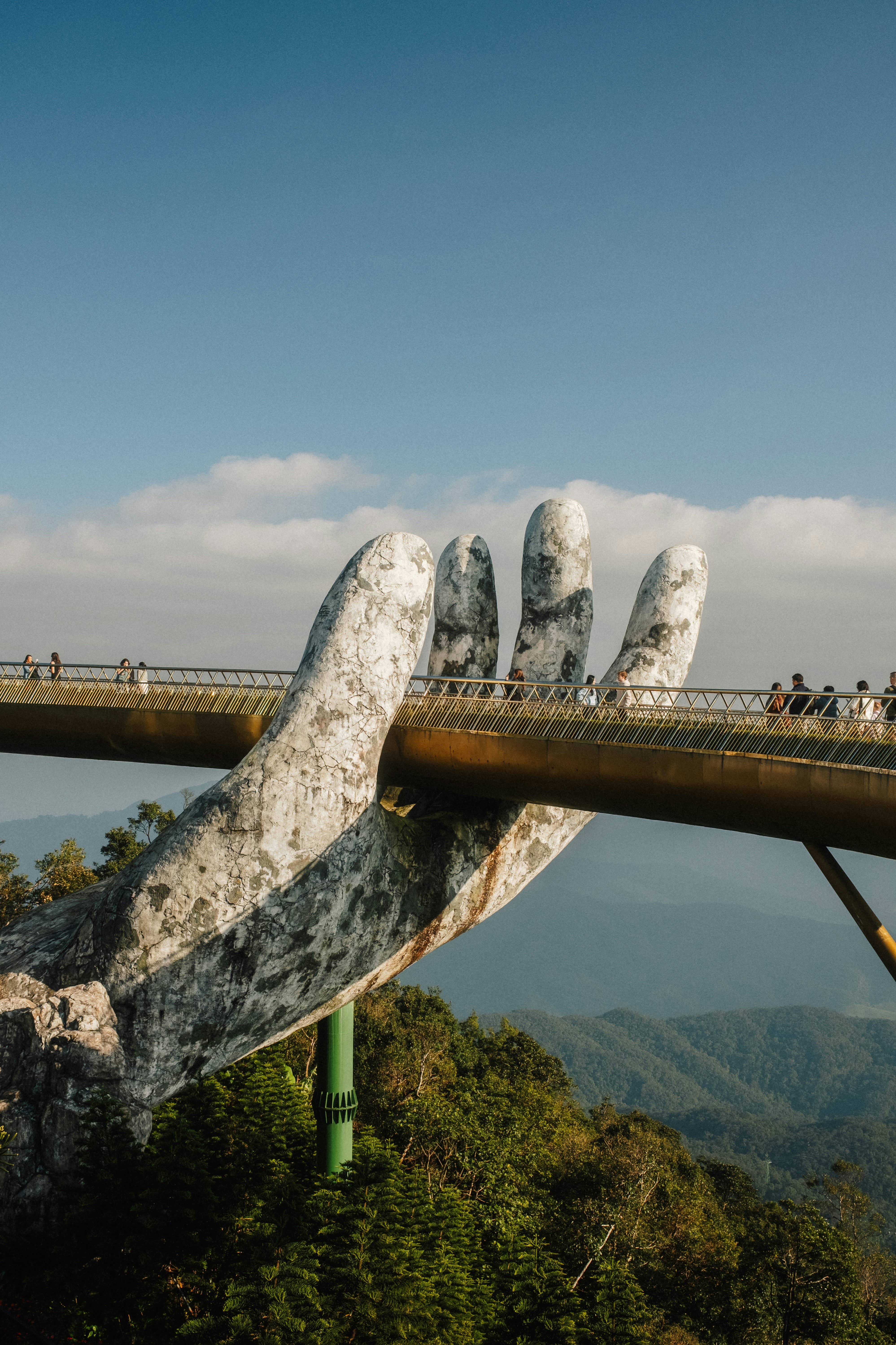 Vietnam's Golden Bridge supported by two giant stone hands, with visitors walking across above forested mountains.