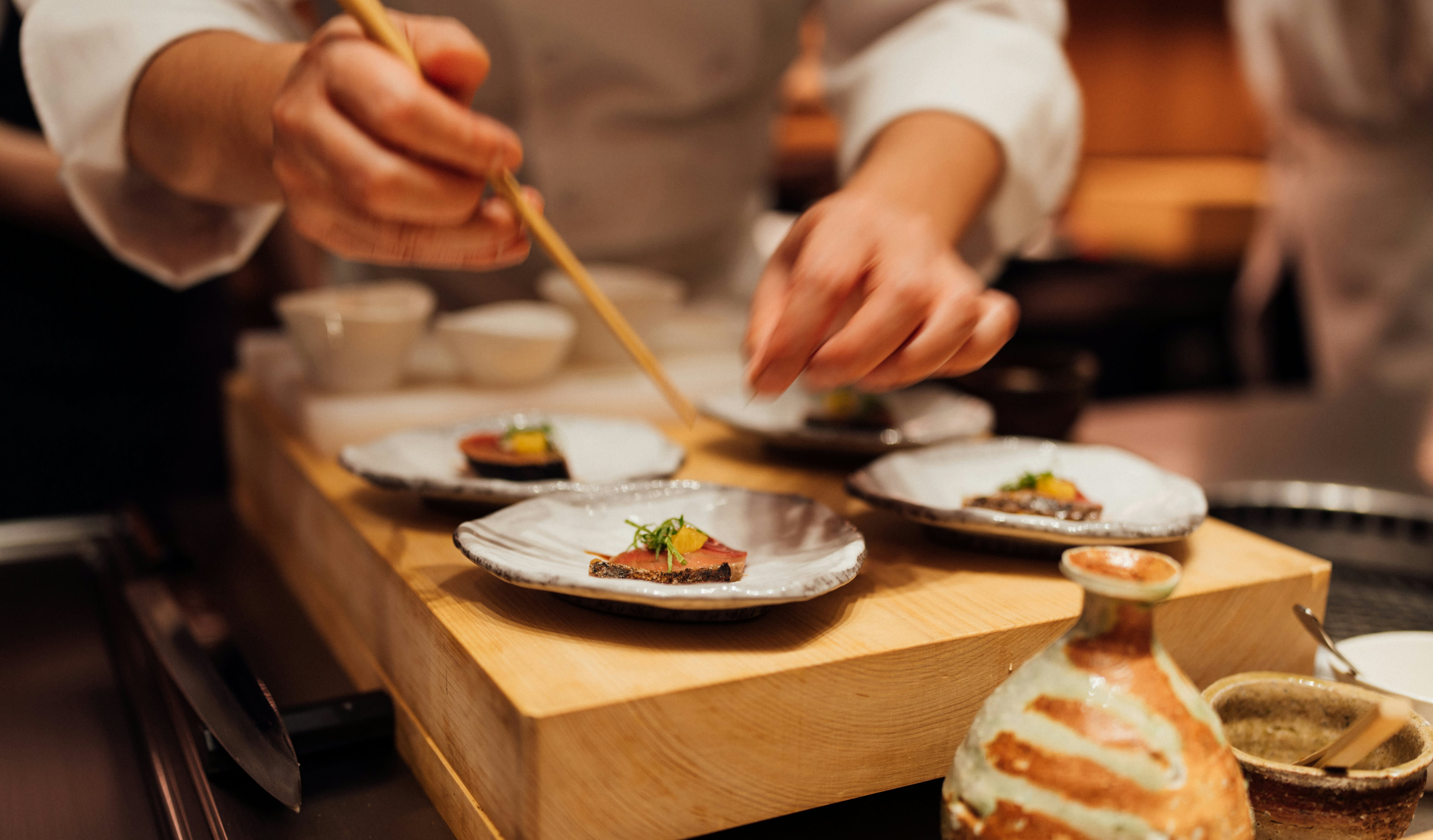 Chef plating elegant Japanese dishes on small ceramic plates atop a wooden board, using chopsticks for precise garnishing.