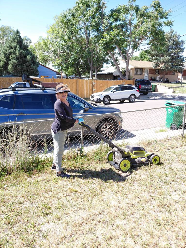 Smiling woman wearing a visor and gloves mowing a dry grass yard with a green electric lawn mower during a volunteer day.
