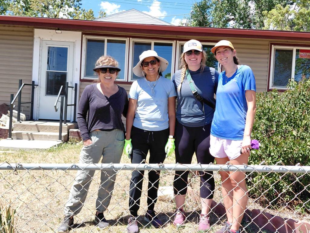 Four smiling women in hats and sunglasses stand behind a chain-link fence in front of a house during a volunteer day.