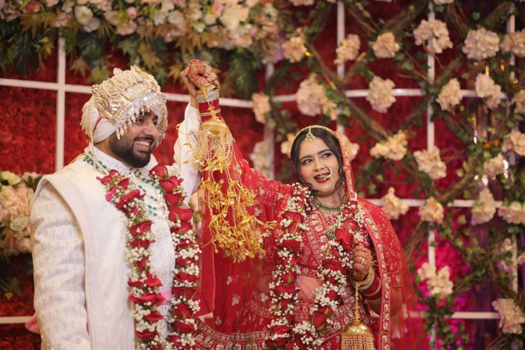 Indian bride in red lehenga and groom in white sherwani raising joined hands joyfully, adorned with floral garlands at a wedding ceremony.