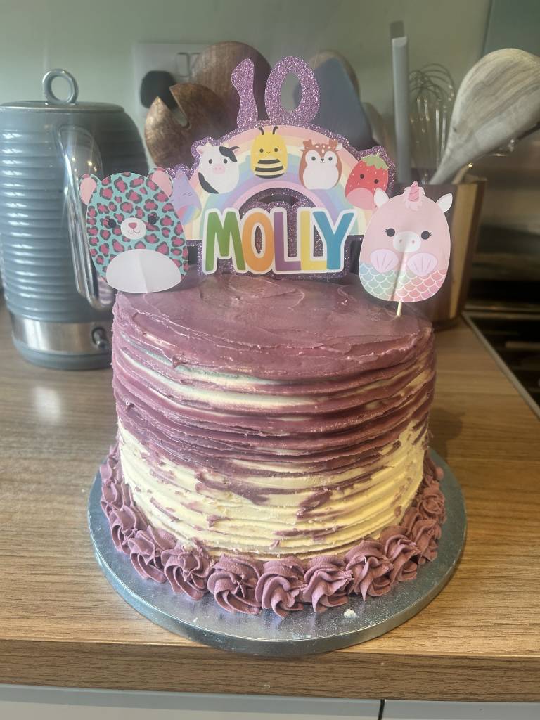 Purple and cream layered birthday cake with Squishmallows topper reading "Molly" and "10," decorated with piped rosettes at the base.