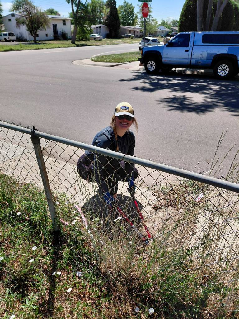 Smiling volunteer in a cap and sunglasses crouching by a chain-link fence, picking up litter along a neighborhood street.