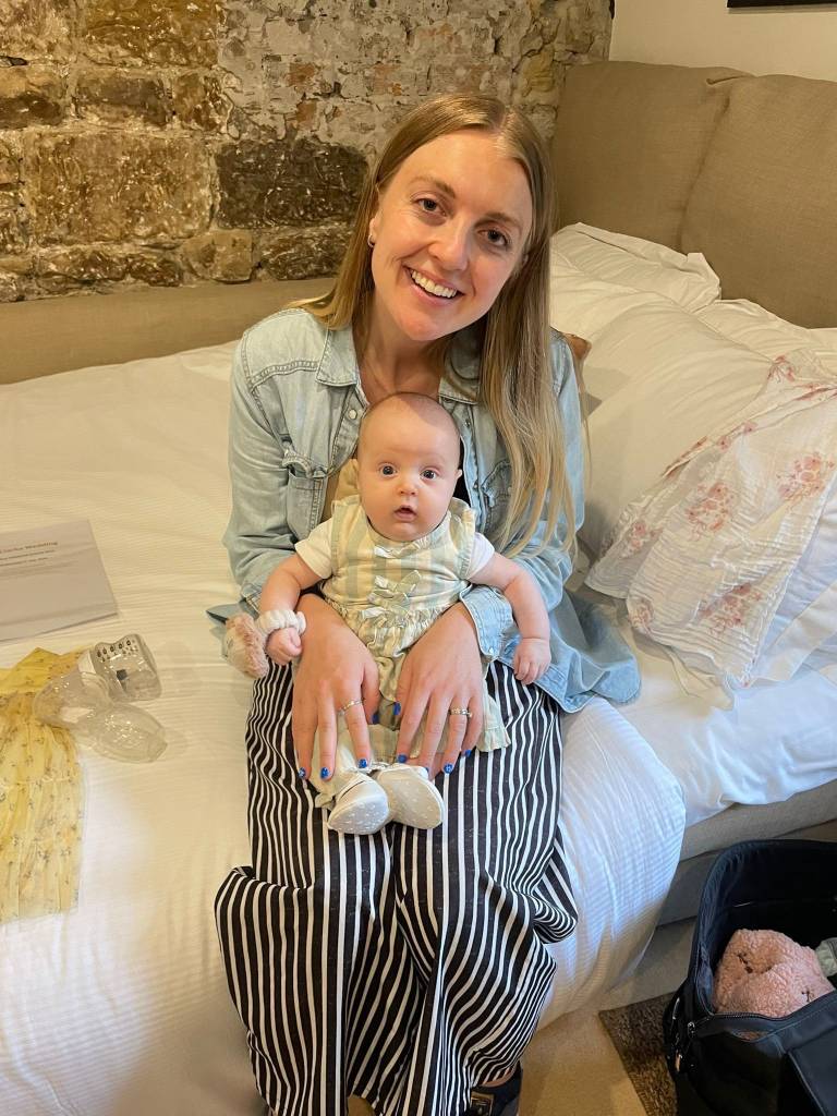 Smiling woman in a denim shirt and striped trousers sitting on a bed, holding a wide-eyed baby in a ruffled outfit.
