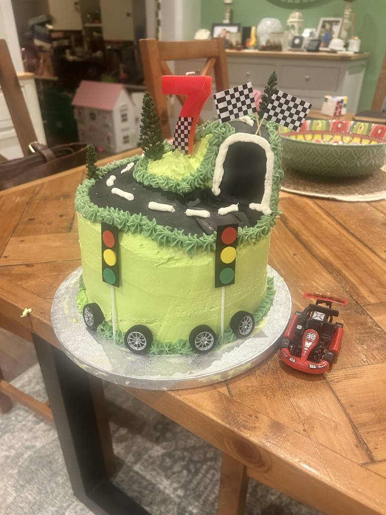 Racing-themed 7th birthday cake with a track, tunnel, checkered flags, traffic lights, wheels, and a red toy race car beside it.