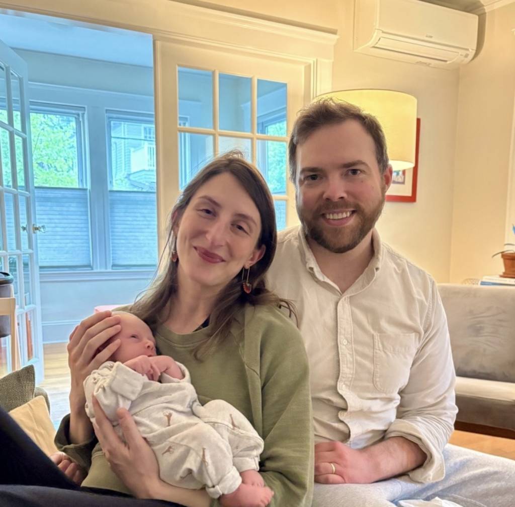Smiling couple sitting together indoors, woman holding a newborn baby dressed in white.