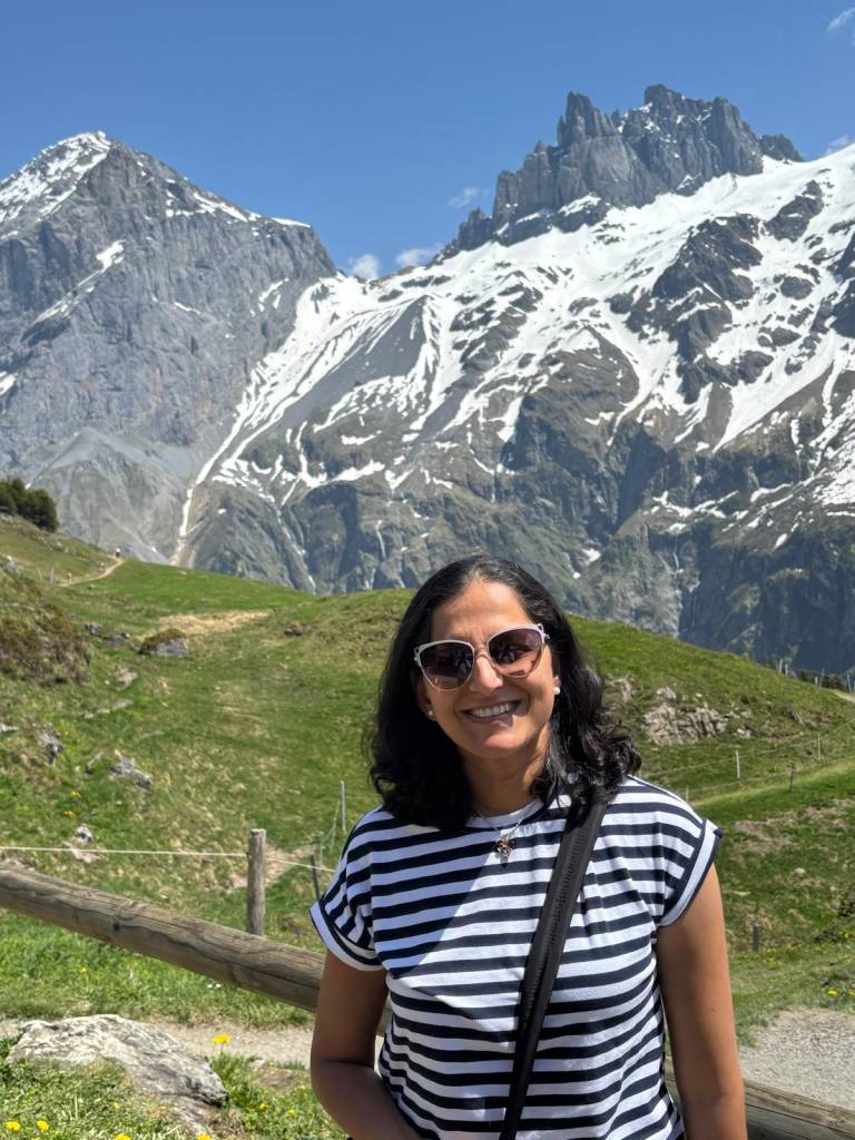Vineeta smiling outdoors in sunglasses and a striped shirt, with snow-capped alpine mountains and green meadows behind her.