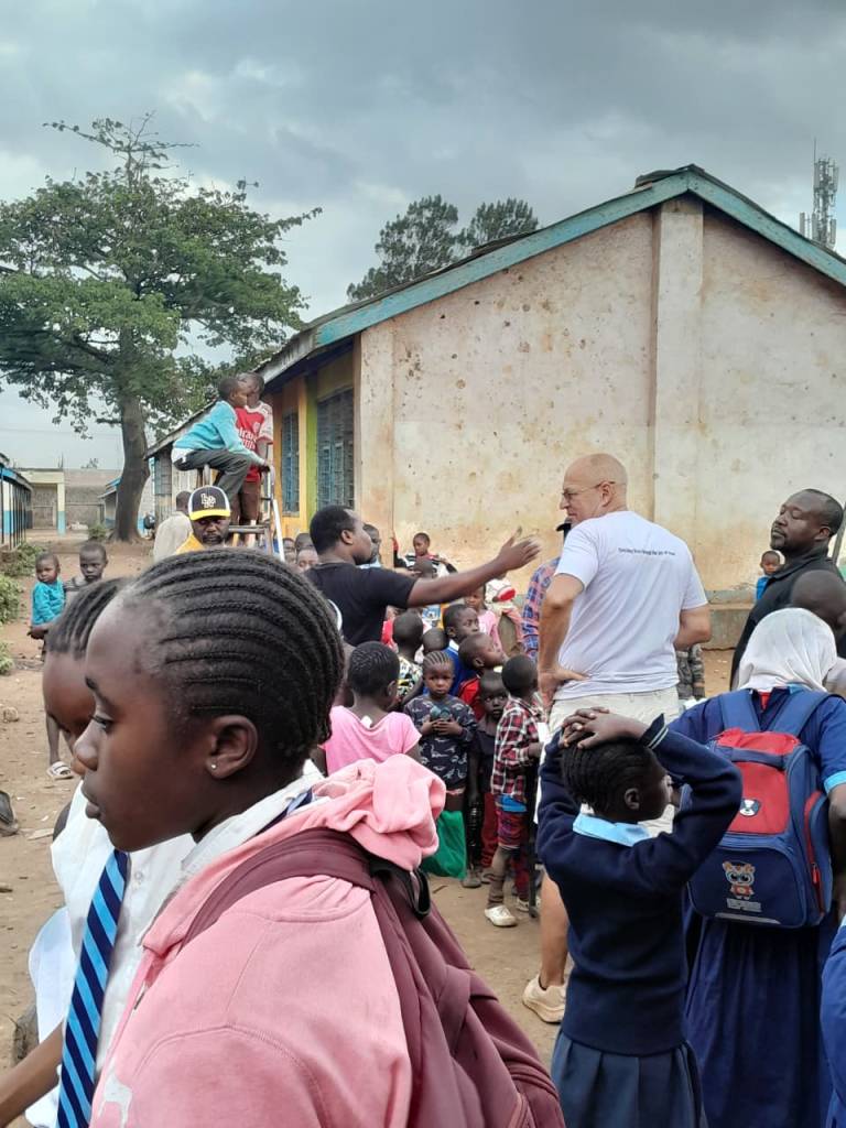 Volunteer in white shirt engaging with a large group of children and adults outside a school building in Africa.