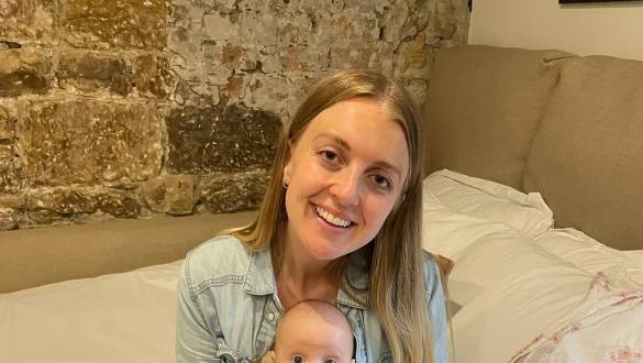 Smiling woman in a denim shirt and striped trousers sitting on a bed, holding a wide-eyed baby in a ruffled outfit.