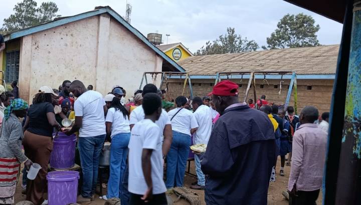 Volunteers in white t-shirts serving food from large buckets to a crowd outside a school building in Africa.