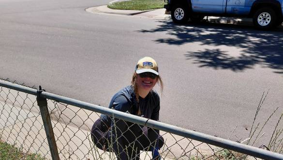 Smiling volunteer in a cap and sunglasses crouching by a chain-link fence, picking up litter along a neighborhood street.