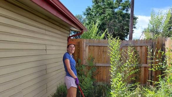 Smiling volunteer in blue shirt and cap holding a rake, clearing overgrown weeds beside a house on a sunny day.