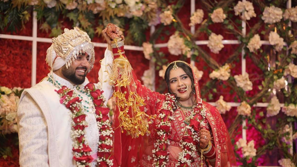 Indian bride in red lehenga and groom in white sherwani raising joined hands joyfully, adorned with floral garlands at a wedding ceremony.