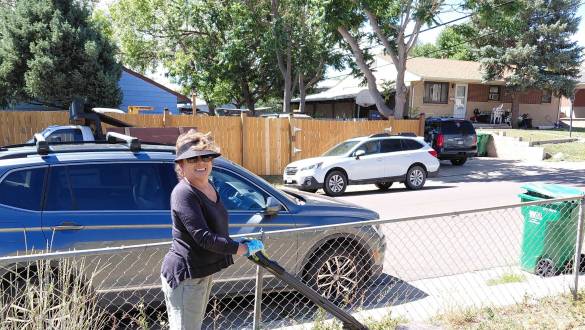 Smiling woman wearing a visor and gloves mowing a dry grass yard with a green electric lawn mower during a volunteer day.