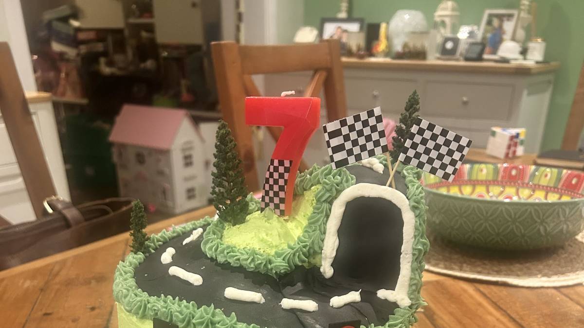 Racing-themed 7th birthday cake with a track, tunnel, checkered flags, traffic lights, wheels, and a red toy race car beside it.