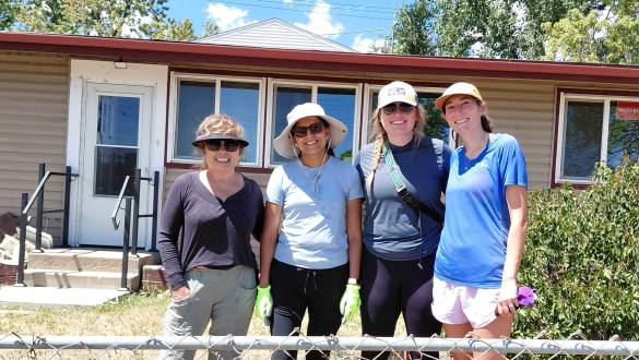 Four smiling women in hats and sunglasses stand behind a chain-link fence in front of a house during a volunteer day.