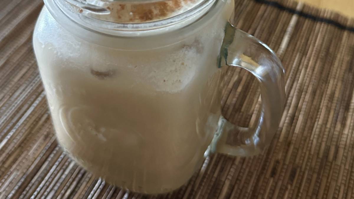 Creamy protein shake in a glass mason jar mug, topped with cinnamon and a rose gold metal straw, on a woven placemat.