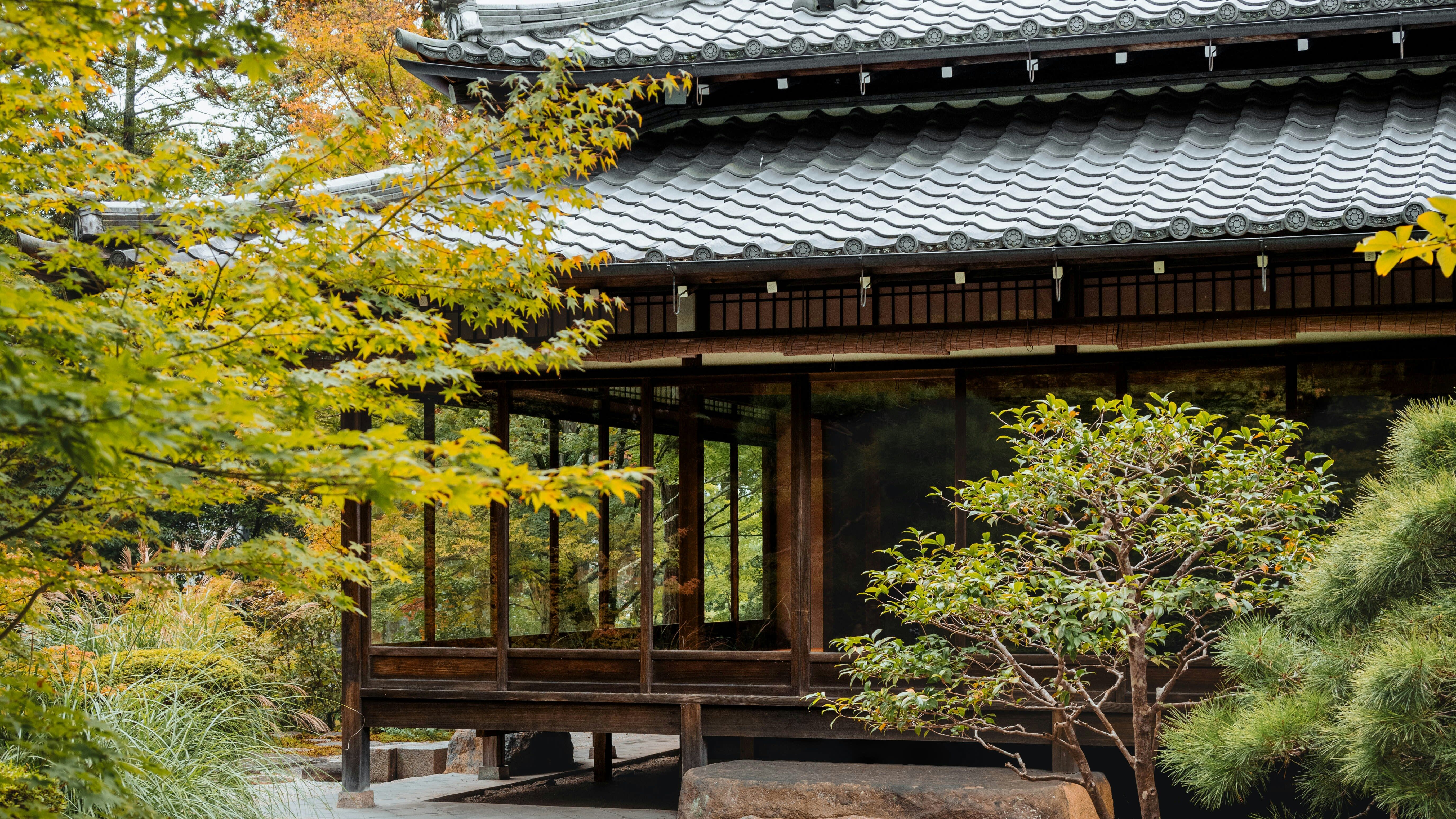 Traditional Japanese pavilion with curved tiled roof, surrounded by autumn maples, pruned trees, and a raked gravel garden.