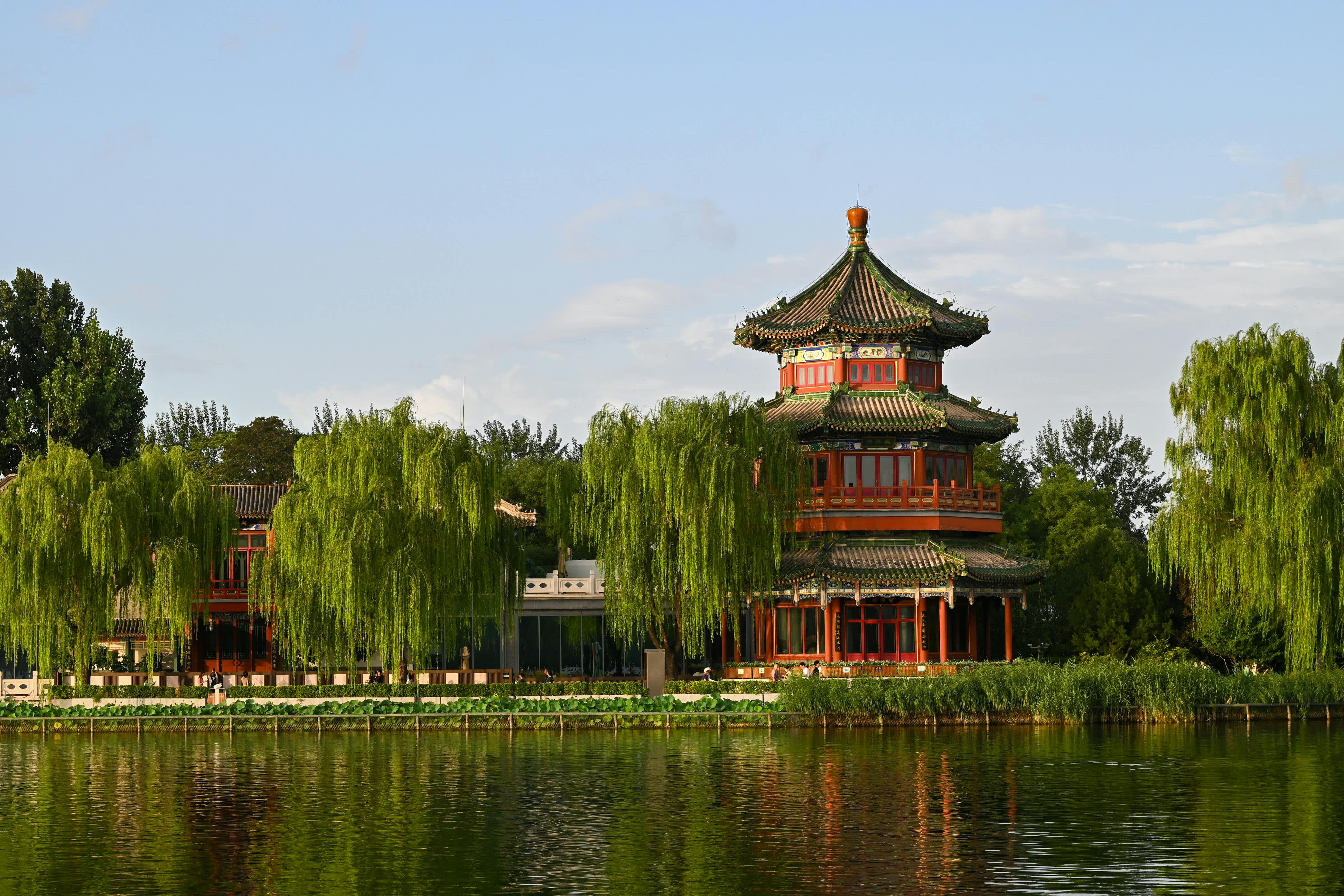 Traditional Chinese pagoda with red and green tiered roofs beside a calm lake, surrounded by weeping willows and lush trees.