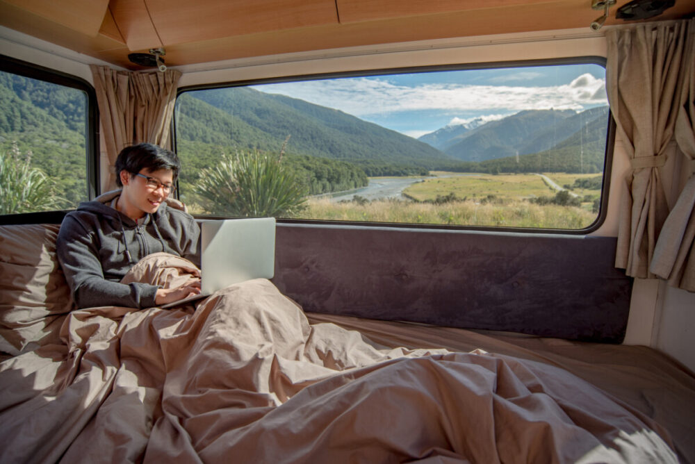 Young Asian man working with laptop computer on the bed in camper van with snow mountain scenic view through the window, digital nomad concept