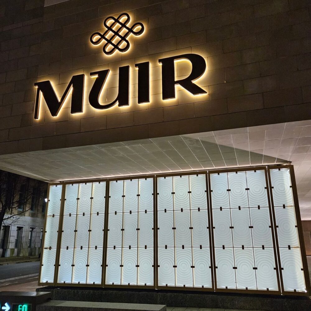 Muir Hotel sign lit from behind. Beside it large wall connected with clips for it is made of Lighthouse lenses.